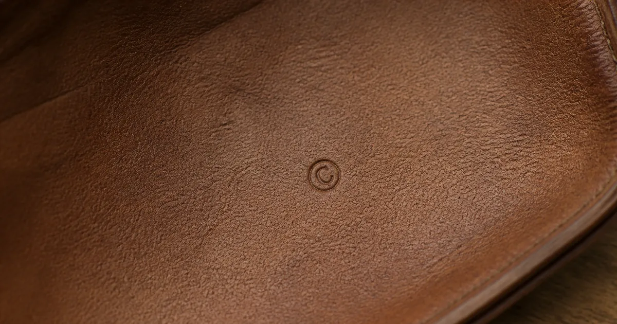 Primo piano di un piccolo marchio a secco impresso sulla pelle interna di una borsa di lusso vintage, allineamento preciso, lettere e numeri nitidi, su una scrivania in rovere opaco in luce naturale morbida (illustrazione generata da IA)