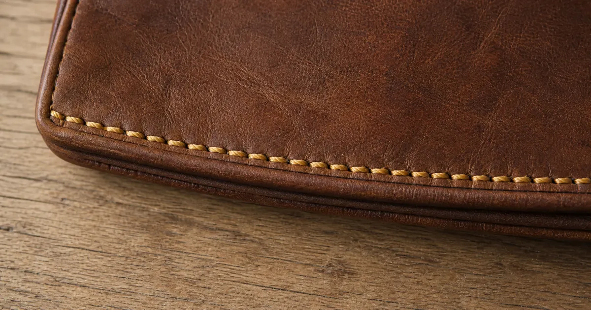 Primo piano della cucitura lungo il bordo di una borsa in pelle vintage, punti fitti e regolari in filo leggermente senape, tutti paralleli al bordo della pelle, su una scrivania in rovere opaco in luce naturale morbida (illustrazione generata da IA)